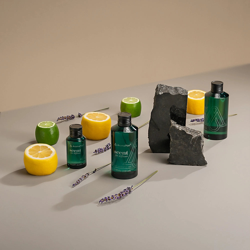 An arrangement of two bottles of fragrance oil placed next to yellow and green citrus fruits and lavender sprigs, with two large rocks in the background.