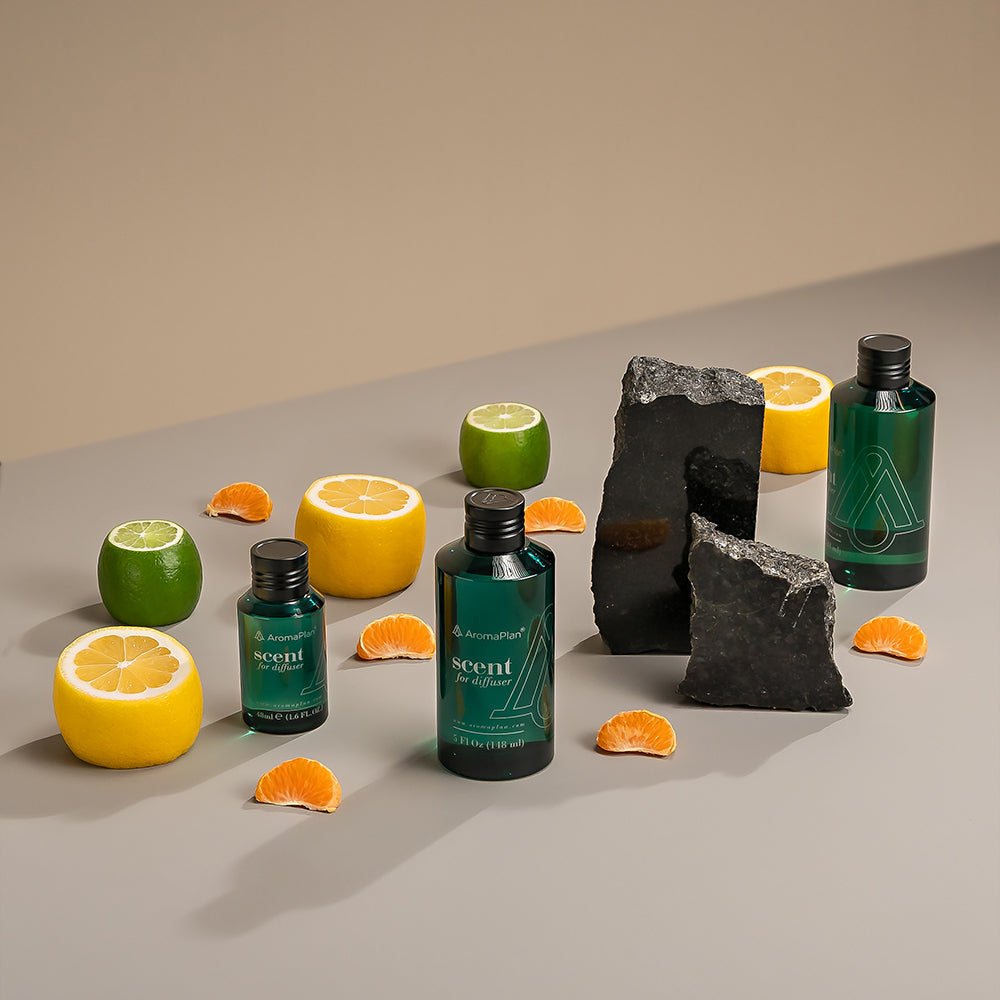 A set of AromaPlan scent diffuser bottles arranged on a neutral-colored surface. The bottles are surrounded by lime slices, pinecones, pine sprigs, and marble blocks, creating a fresh and natural aesthetic.
