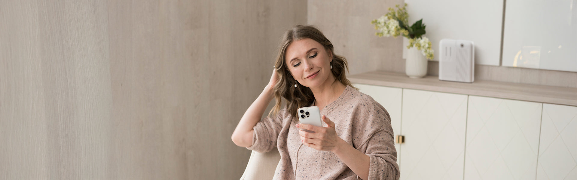 A woman with light brown hair, wearing a beige sweater, is sitting and holding a phone while smiling contently in a modern living space with a soft-focus background.
