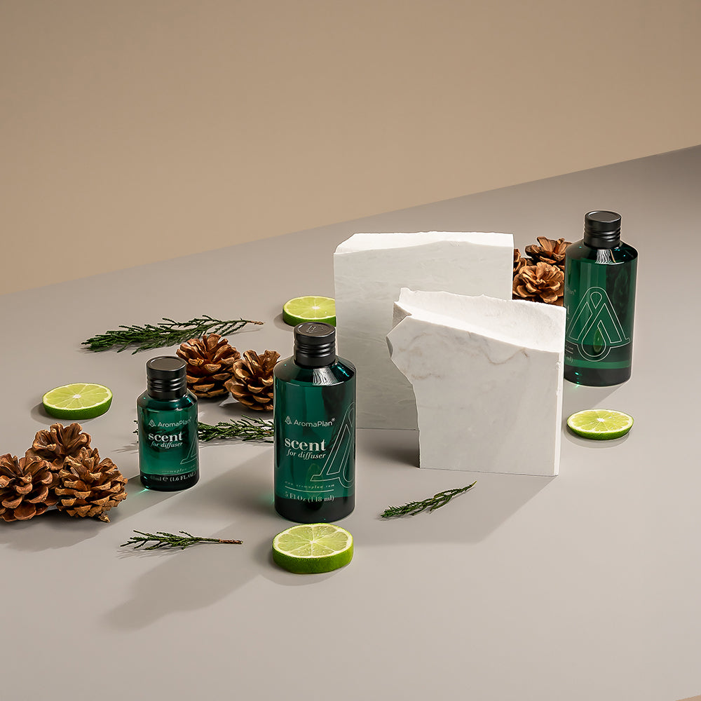 A set of AromaPlan scent diffuser bottles arranged on a neutral-colored surface. The bottles are surrounded by lime slices, pinecones, pine sprigs, and marble blocks, creating a fresh and natural aesthetic.