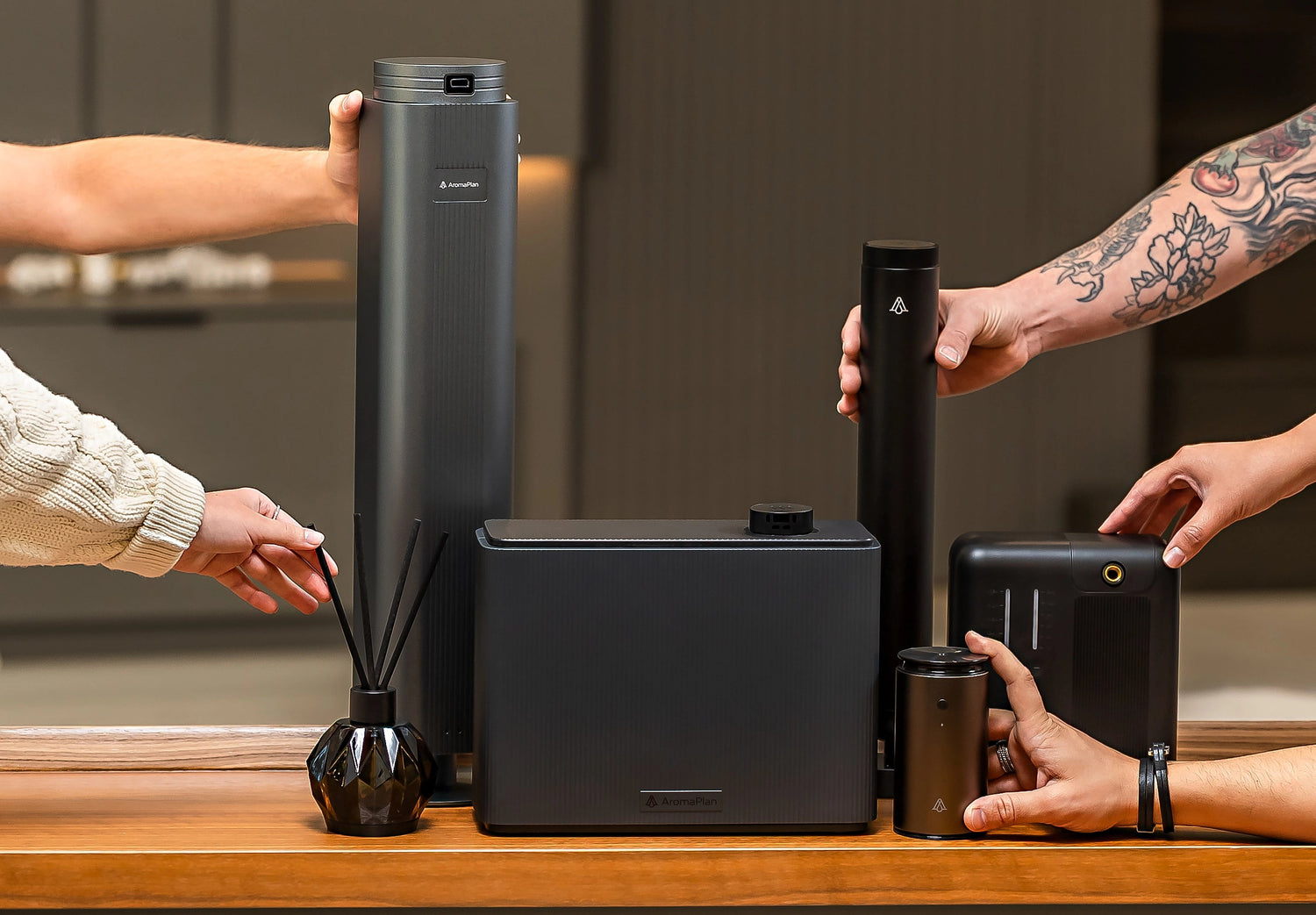 Various black scent diffusers displayed on a wooden surface, with multiple hands reaching to interact with them. The diffusers vary in size and shape, with a mix of cylindrical and rectangular designs.